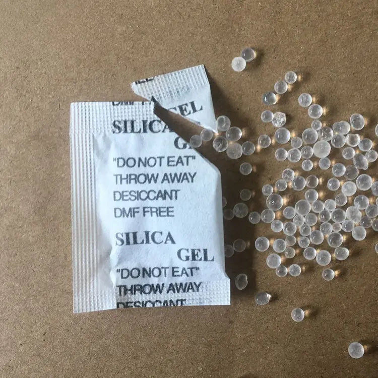 Open silica‑gel desiccant packet with clear moisture‑absorbing beads spilled on a cardboard surface